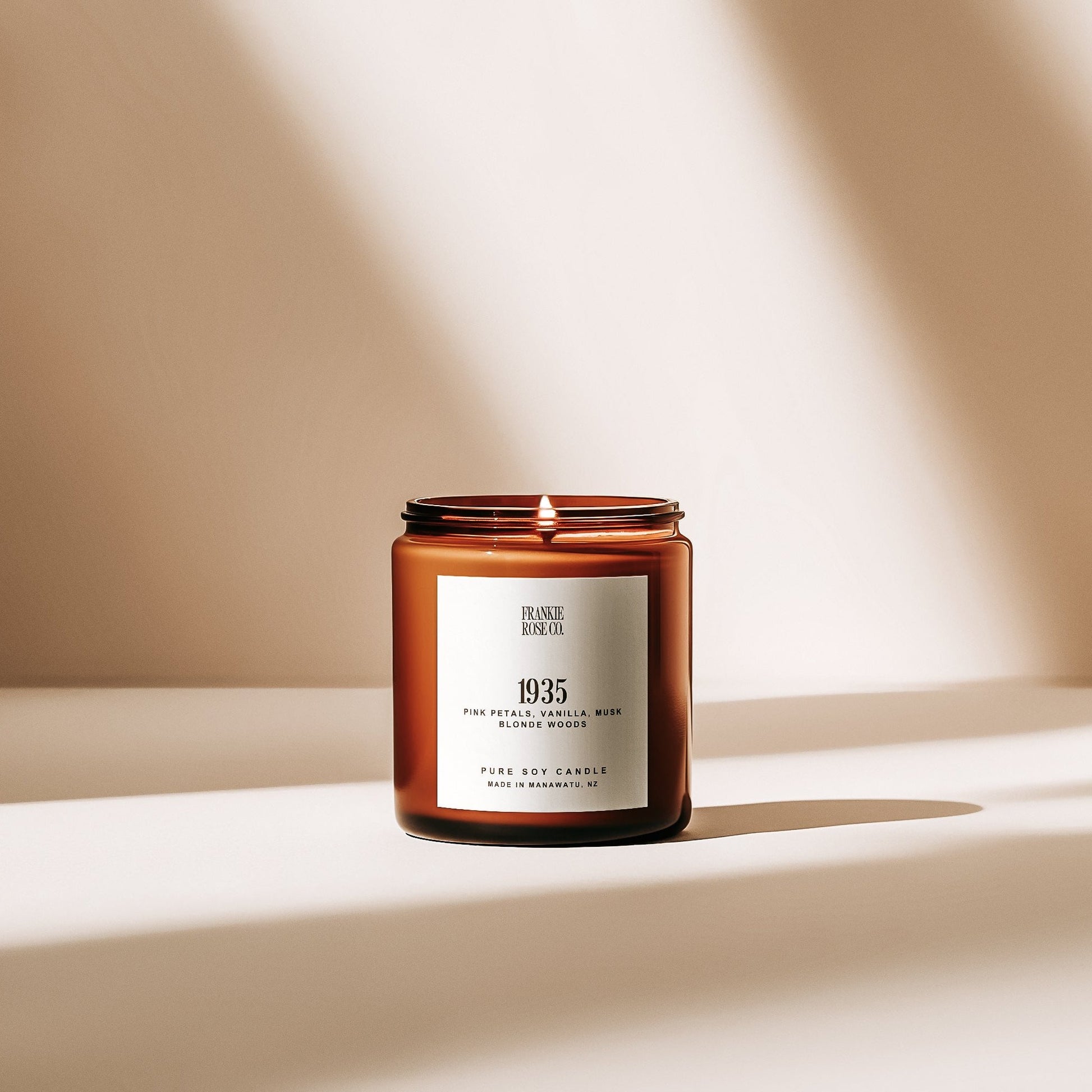 A beautifully lit candle labeled '1935,' with a soft, warm glow that captures the essence of its sweet and floral fragrance. The delicate scent of pink chiffon and bergamot rises gently, followed by the romantic heart notes of jasmine, rose, and vanilla. A base of sandalwood and musk provides a warm, inviting finish, perfect for creating a relaxing and romantic atmosphere in any space."